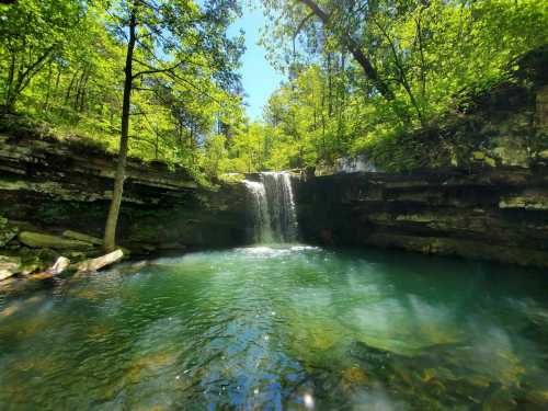 A serene waterfall cascades into a clear pool, surrounded by lush green trees and rocky cliffs under a bright blue sky.