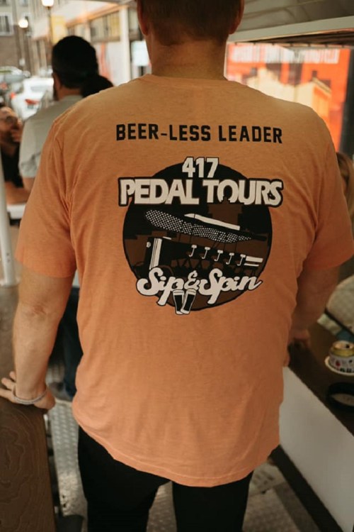 A person wearing an orange t-shirt with "Beer-Less Leader" and "417 Pedal Tours" printed on the back.
