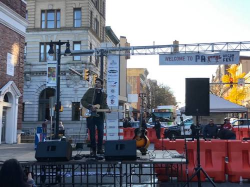 A musician performs on stage at a street festival, with buildings and a crowd in the background.