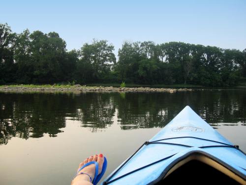 A view from a kayak, showing a foot in a sandal and calm water reflecting trees on the shore.