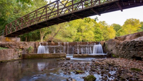 A wooden bridge spans a serene stream with gentle waterfalls, surrounded by lush greenery and rocky banks.