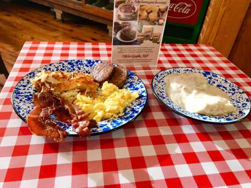 A blue and white plate with scrambled eggs, bacon, sausage, and hash browns next to a bowl of gravy on a checkered tablecloth.