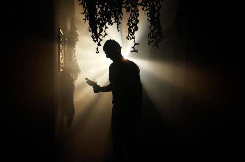 A silhouetted figure stands in a dimly lit space, with beams of light shining through, creating a mysterious atmosphere.