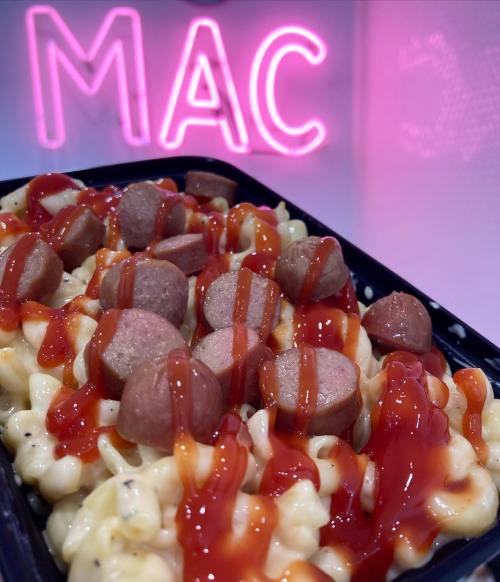 A tray of macaroni and cheese topped with sliced hot dogs and drizzled with ketchup, with a neon "MAC" sign in the background.