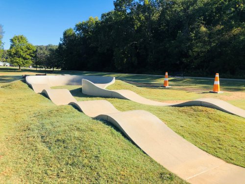 A winding concrete pump track surrounded by grass and trees, with traffic cones marking the area.