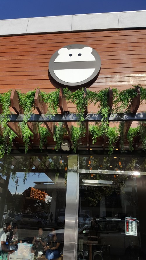 A storefront with a smiling bear logo and greenery hanging from a wooden awning. People are visible inside.