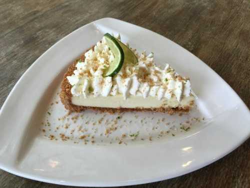 A slice of key lime pie topped with whipped cream and a lime wedge, served on a white plate with a graham cracker crust.