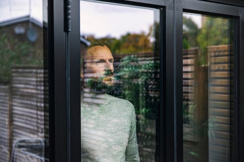 A man gazes thoughtfully out of a large window, reflecting on the greenery outside.