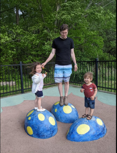 A man stands on a blue, polka-dotted structure in a playground, with two children beside him, all smiling and playing.