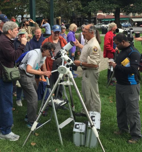 A group of people gather around a telescope, engaging with an expert in a park setting.