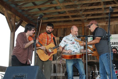 Four musicians perform on stage, playing a fiddle, guitar, banjo, and bass in a rustic wooden setting.
