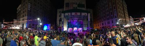 A large crowd gathers at night for a concert, with a stage illuminated by colorful lights in a city setting.