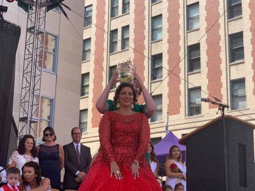 A woman in a red gown smiles as another places a crown on her head during a celebration on stage.