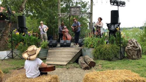 A band performs on stage at Old Timers Day, with a child playing guitar in the foreground and a lush garden backdrop.