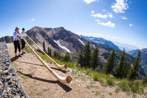 Two musicians in traditional attire play long wooden horns on a mountain peak, surrounded by scenic views and trees.