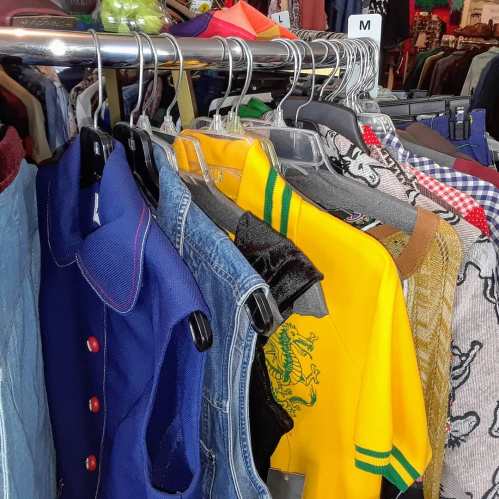 A colorful assortment of clothing hangs on hangers, including shirts, jackets, and a denim vest.