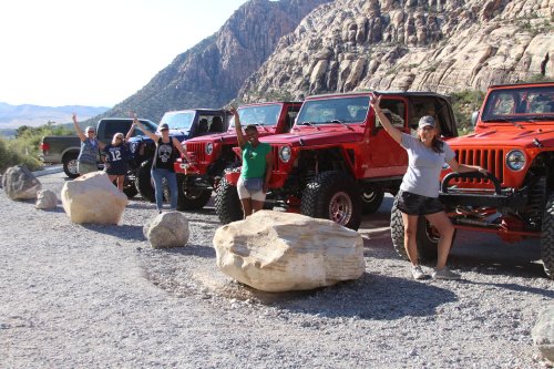 A group of five people poses beside red off-road vehicles in a rocky landscape, raising their arms in celebration.