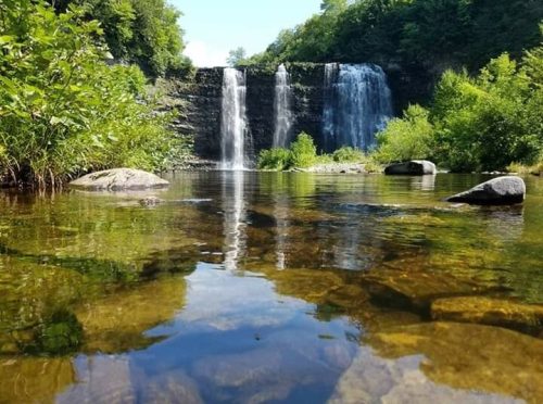 A serene waterfall cascades into a clear pool, surrounded by lush greenery and rocky terrain under a bright blue sky.