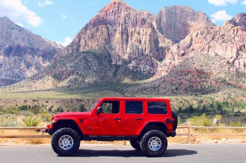 A red Jeep parked on a road with stunning mountains in the background under a clear blue sky.