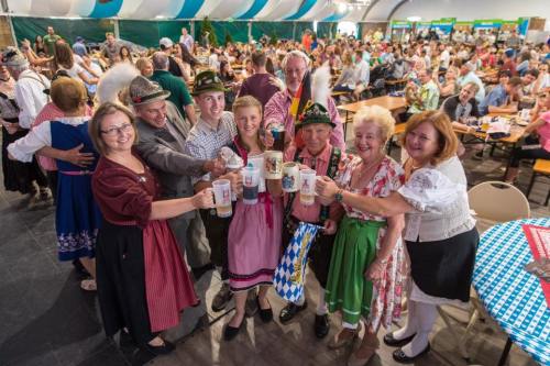 A group of people in traditional attire holding beer mugs, smiling at a festive event with a large crowd in the background.