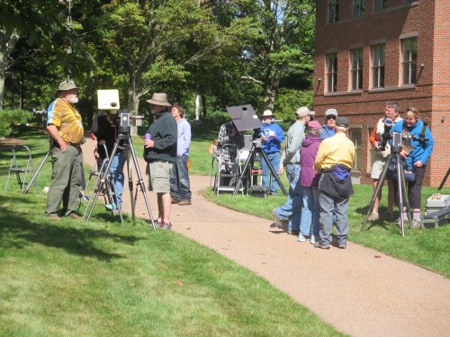 A group of people set up equipment outdoors, with tripods and cameras, near a brick building and green trees.