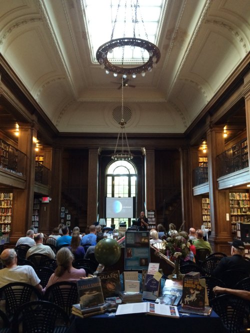 A library interior with a chandelier, bookshelves, and an audience watching a presentation on a screen.