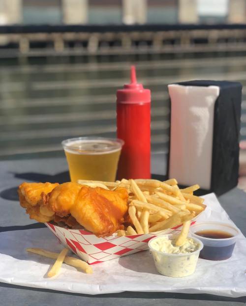 A basket of golden fried fish and fries, with dipping sauces, a beer, and ketchup on a table by the water.