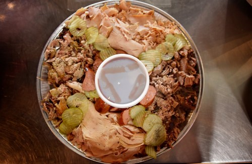 A platter of assorted meats, including turkey and pulled pork, garnished with pickles and a small cup of sauce in the center.