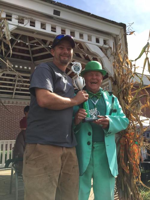 Two men pose together, one in a green suit holding a trophy, celebrating an award at an outdoor event.