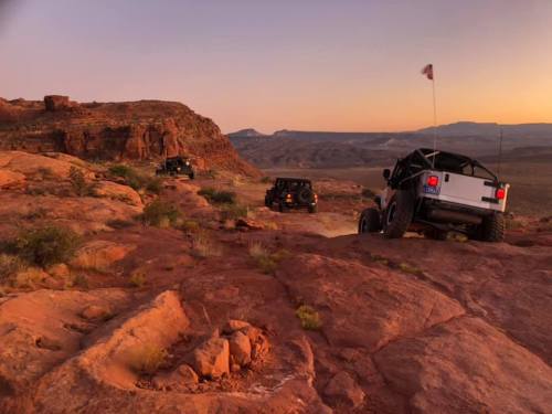 Off-road vehicles navigate rocky terrain at sunset, with a vast desert landscape in the background.