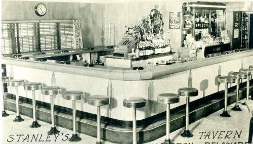 A vintage bar interior with a curved counter, stools, and shelves of bottles, labeled "Stanley's Tavern."