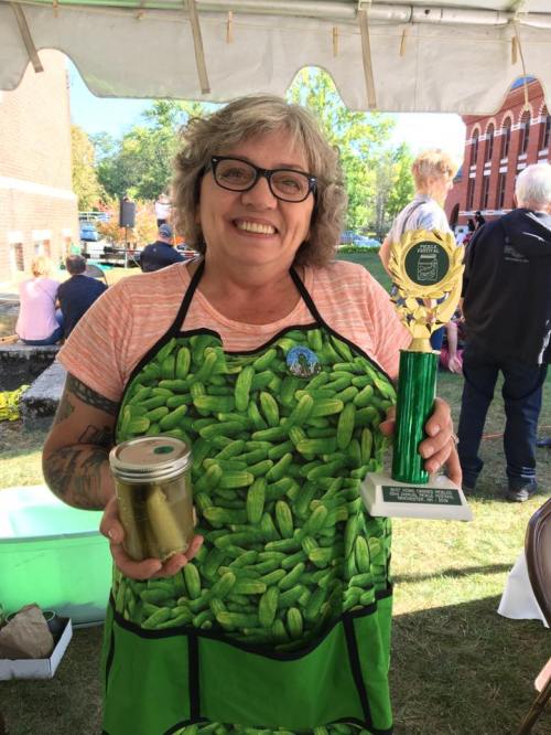 A smiling woman in a pickle-themed apron holds a jar of pickles and a trophy at an outdoor event.