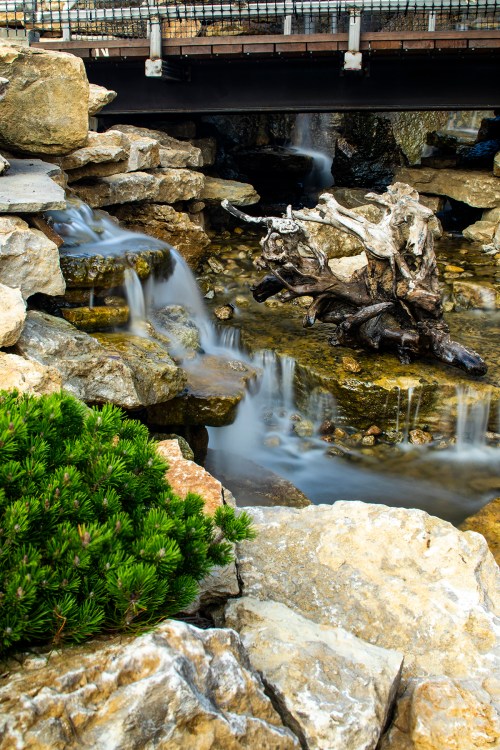A serene water feature with flowing water over rocks, surrounded by greenery and a wooden bridge above.