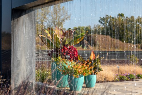 A serene scene featuring colorful potted plants behind a water curtain, with a blurred natural backdrop.