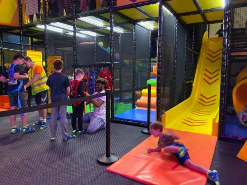 Children play in a colorful indoor trampoline park, with slides and safety barriers, while adults supervise.