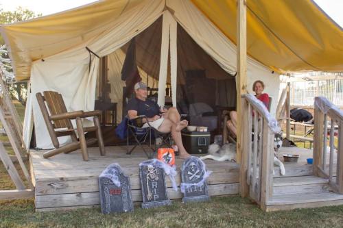 A tent with two people sitting outside, decorated with Halloween-themed gravestones and a dog nearby.