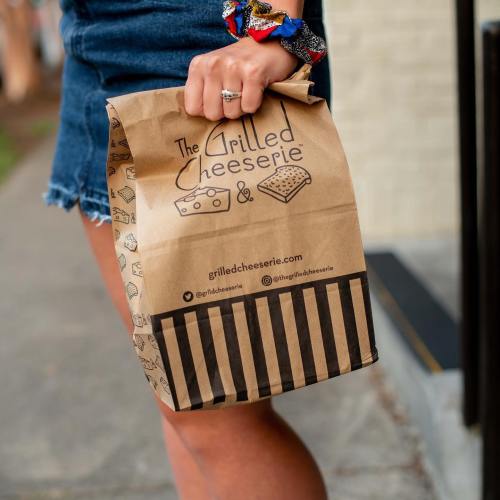 A person holds a takeout bag from The Grilled Cheeserie, wearing a blue top and denim shorts.