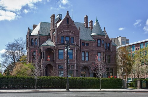 A large, historic mansion with a stone facade and green roof, surrounded by trees and a fence, under a clear blue sky.