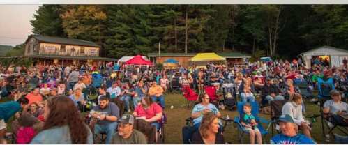 A large crowd gathers outdoors at a festival, seated in chairs with tents and trees in the background.