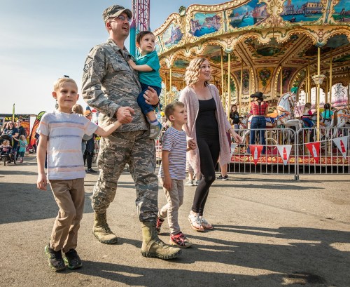 A military parent walks with three children near a carousel at a fair, enjoying a sunny day together.
