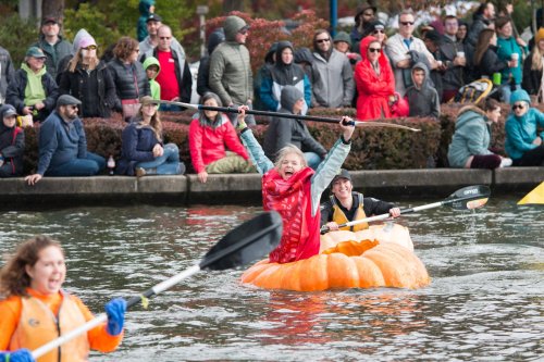 A person in a pumpkin kayak celebrates while paddling, surrounded by a cheering crowd along the water's edge.