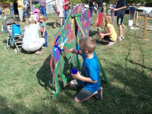 A child paints a colorful horse cutout at an outdoor event, surrounded by other children and adults.