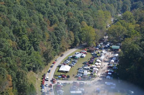 Aerial view of a festival with tents and cars along a winding road, surrounded by lush green trees.