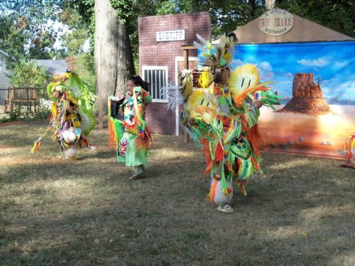 Three dancers in colorful traditional costumes perform outdoors, surrounded by trees and a backdrop of a desert landscape.