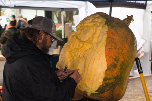A person carves a detailed design into a large pumpkin at an outdoor market.