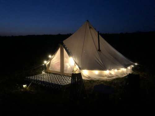 A lit tent with string lights at dusk, set in a grassy field, creating a cozy outdoor atmosphere.