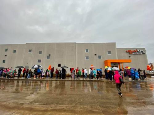 A long line of people with umbrellas waiting outside a gray building on a rainy day.