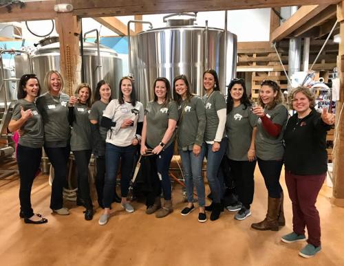 A group of ten women in matching gray shirts pose together in a brewery, smiling and holding drinks.