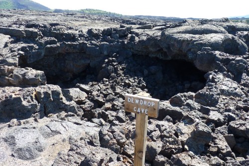 Sign for Dewdrop Cave in a rocky, volcanic landscape with a cave entrance visible in the background.