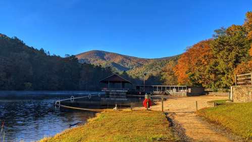 A serene lake scene with a wooden dock, surrounded by autumn trees and mountains under a clear blue sky.